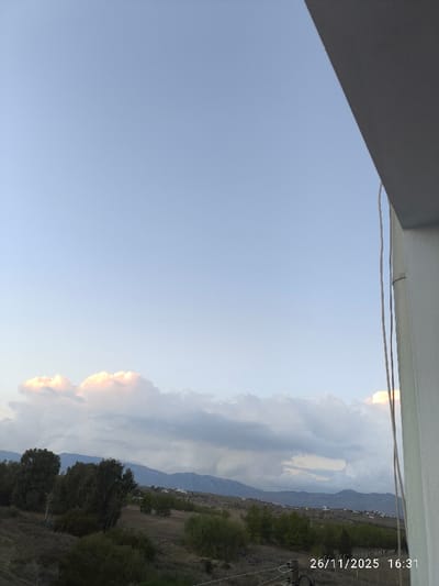The image presents a panoramic landscape view from an elevated position in North Nicosia, Northern Cyprus / Cyprus, recorded on 26/11/2025 at 16:31. The upper approximately 60% of the frame features a pale blue sky, transitioning to lighter tones towards the horizon. A band of cumulus clouds stretches across the mid-ground, with some clouds on the left exhibiting distinct pinkish-orange illumination from sunlight, contrasting with predominantly gray clouds. In the background, a range of hazy, blue-gray mountains is visible. The foreground and mid-ground consist of arid, undulating terrain covered in sparse, brownish-green scrubland and scattered trees, including taller, darker green specimens and smaller, lighter green bushes. Distant residential structures are faintly discernible on the mountain slopes. No people are present. A white architectural element, consistent with a building edge or balcony, occupies the far right side of the image, extending diagonally downwards from the top right corner, with two thin, vertical white wires or cables running alongside it.