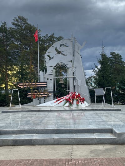 The image shows a war memorial in Belorechensky, Russia. The memorial is constructed of light gray granite and white metal. The central design element is a large metal cutout depicting stylized birds, a window, and the numbers "1941-1945." Red and white flowers are placed at the base of the memorial. A red flag flies to the side. Behind the memorial are evergreen trees. The weather appears cloudy. The memorial is elevated, with granite steps leading up to it. A small plaque is visible on a stand to the right of the main structure. The background includes glimpses of other buildings and structures. The image appears to have been taken at ground level.
