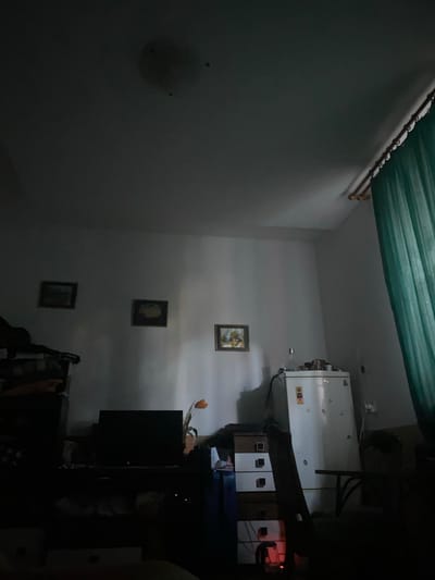 The image depicts a dimly lit room, possibly a bedroom or living area, with a predominantly gray and white color scheme. The room contains a white refrigerator on the right side, a chest of drawers with multiple compartments, a dark television set with a red indicator light, a single orange flower in a vase, and three framed pictures hanging on the wall. A green curtain is visible on the far right. The lighting is low, casting shadows throughout the room. No individuals are present in the scene. The overall impression is one of a functional but somewhat cluttered living space. It is not possible to determine the specific location within the city of Zgierz, Poland from the visual information provided.