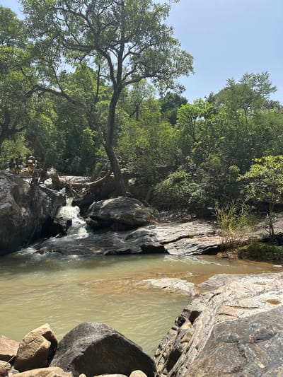 The image depicts a natural scene in Fobur, Nigeria, featuring a small waterfall cascading over rocks into a body of water. Lush green trees and foliage dominate the left side of the frame, with their branches extending towards the bright, clear sky. Large, textured rocks line the right side and surround the waterfall, creating a rugged and natural environment.
The water appears somewhat murky, with a greenish-brown hue. The sunlight suggests it is daytime, likely mid-morning or afternoon, given the shadows and the brightness of the sky. The overall impression is one of a serene, somewhat wild, natural landscape. There are no discernible people or specific objects of interest, other than the natural elements of the waterfall, rocks, trees, and water. The focus is entirely on the natural beauty of the location. The scene evokes a sense of tranquility and the raw beauty of nature. No text is visible in the image.
