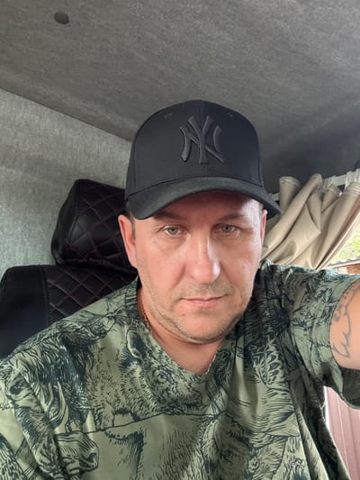 A man wearing a black baseball cap with a New York Yankees logo and a camouflage t-shirt is sitting in what appears to be the cab of a vehicle. The background shows a textured grey ceiling, a black leather seat with red stitching, and beige curtains. A portion of the man's arm with a tattoo is visible on the right side of the image. The lighting suggests it is daytime. The location is not specified in the image itself, but context suggests Yuzha, Russia.