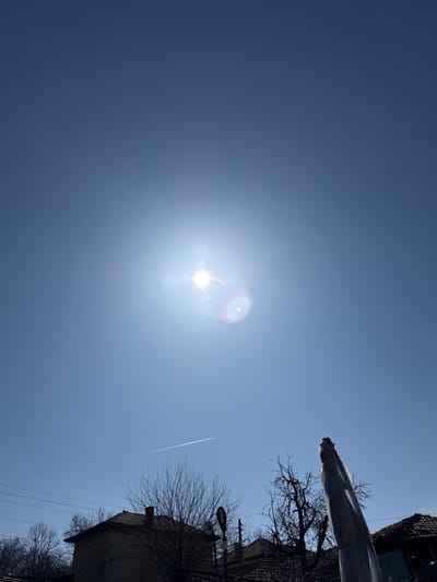 The scene depicts a sunny daytime view in Devetaki, Bulgaria. The upper two-thirds of the image show a clear, light blue sky dominated by the bright, glaring sun in the upper-center, producing a prominent lens flare with visible green and purple chromatic aberrations. A thin, white aircraft contrail extends horizontally across the middle-left sky. The lower portion of the image reveals fragmented architectural and natural elements. Multiple buildings with traditional red-tiled roofs are visible, accompanied by numerous bare deciduous trees, indicating a leafless season. Power lines traverse the mid-left, and a black street light is present near the center-left. On the far right, a tall, vertical object is partially obscured by white or translucent protective covering, tapering upwards from the lower right corner. No individuals are present.
