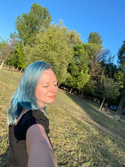 A young woman with bright blue hair is taking a selfie outdoors on a sunny day. She is wearing a black top and her eyes are closed, with a relaxed expression on her face. Her skin is glowing in the sunlight. Behind her, the scene is a grassy field with trees and a clear blue sky. In the background, a dark-colored car and some trees are visible. The sunlight suggests it is either morning or late afternoon. The overall mood of the image is peaceful and serene.