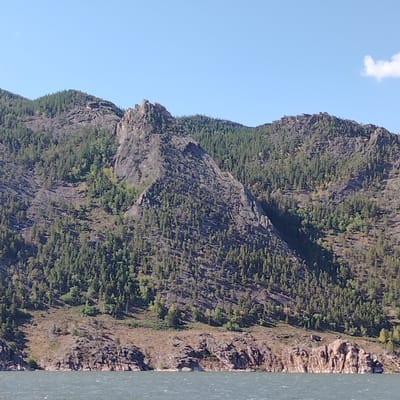 This is a wide-angle, landscape photograph taken on a clear, sunny day from the perspective of a body of water. The image features a large, rugged mountain range covered in dense green pine trees. The mountains are characterized by steep, rocky slopes and jagged peaks. In the foreground, the dark, choppy water reflects the blue sky. The shoreline is rocky, with exposed rock formations meeting the water. To the right of the frame, a small, wispy white cloud is visible in the sky. The scene is natural and undeveloped, suggesting a remote or natural park area within or near the city of Торайғыров, Kazakhstan, known for its mountainous terrain. There are no people or animals visible in the image.