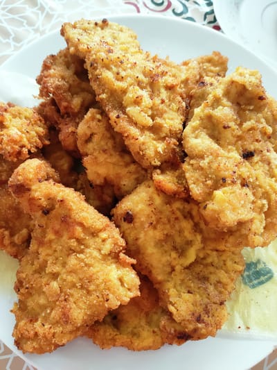 A close-up, overhead shot shows a pile of golden-brown, breaded chicken cutlets on a white plate. The cutlets are irregularly shaped and appear to be fried, with a crispy, textured surface. Some of the cutlets show darker brown spots, indicating they are well-cooked. The plate is placed on a patterned surface, possibly a tablecloth, with warm beige and brown tones and geometric designs. To the right of the plate, a small section of a light yellow paper napkin with a green emblem is visible. The lighting suggests an indoor setting, possibly a home or restaurant. The scene is devoid of any people or specific landmarks that would indicate a location in the city of Ariana, Tunisia.