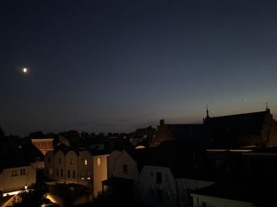 The image depicts a nighttime cityscape, likely taken from an elevated vantage point, overlooking a collection of buildings and the sky. The sky is a deep, dark blue, transitioning to a lighter gradient near the horizon. In the upper left quadrant of the sky, a crescent moon is visible, illuminated by an unseen light source. A few faint stars are also scattered across the sky.
The foreground is dominated by the dark silhouettes of buildings. On the left, a row of houses with illuminated windows suggests occupancy. Their architectural style appears to be traditional, with some featuring arched rooflines. Further back and slightly to the right, a larger building with a more elaborate facade, possibly a church or historical structure, stands out with its prominent roofline and illuminated windows. A red building with white-trimmed windows is also visible amongst the darker structures. Trees and vegetation are present, creating darker masses within the cityscape. The overall impression is one of a quiet, residential area at night. The provided crops confirm the presence of the moon, stars, and various building structures. The location is specified as Deventer, Netherlands.