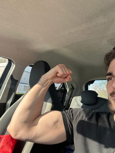 The image shows a man with brown hair and a beard inside a car. He is wearing a black t-shirt and flexing his right arm, which has a tattoo. The interior of the car is gray and black. Through the car windows, buildings can be seen, suggesting a location within Volketswil, Switzerland. The lighting indicates it is daytime.