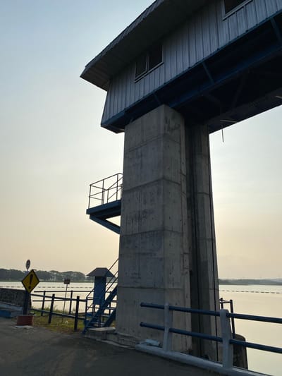 The image shows a concrete structure that appears to be part of a dam or water control gate, situated next to a body of water. The structure has a utilitarian design, with a prominent concrete tower supporting a metal building with windows and a corrugated roof. A metal staircase with a railing leads up to a platform attached to the side of the concrete tower. A blue metal railing lines a walkway in the foreground, and a road is visible to the left, with a yellow diamond-shaped warning sign indicating a hazard and a circular sign with a red bar over a letter 'R'. The sky is a pale, hazy blue, suggesting it might be early morning or late afternoon. Trees and distant structures are visible on the far bank of the water. The scene is devoid of people. This structure is located at the Tuntang Dam in Bandungan, Semarang Regency, Indonesia, which is part of the raw water infrastructure for the city of Semarang.