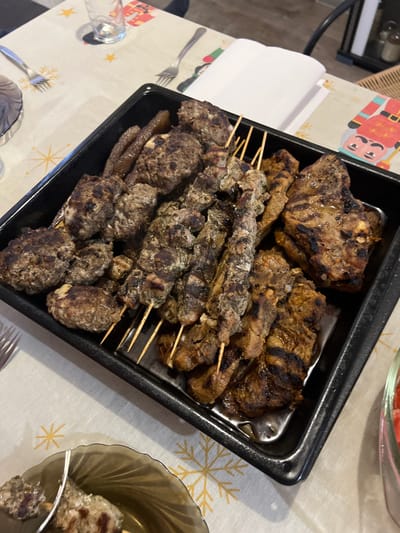 The image displays a black rectangular serving tray, centrally positioned on a dining table, containing a variety of cooked meats. The table is covered with a light-colored tablecloth featuring a pattern of golden snowflakes and festive cartoon figures. The tray holds multiple irregularly shaped grilled meatballs or patties on the left, several wooden skewers with grilled meat pieces in the center, and flatter, broader cuts of grilled meat (likely chops or steaks) on the right, all showing distinct char marks. Some dark, elongated items are present in the upper-left portion of the tray, which also contains accumulated liquid. On the surrounding tablecloth, a clear drinking glass, two silver forks, and a folded white paper napkin are visible. A translucent dark-tinted glass plate holding a single meat skewer is partially visible in the bottom-left, and a red-rimmed dish is partially visible on the far right. No individuals are present in the image. The scene is located in Velichkovo, Bulgaria.