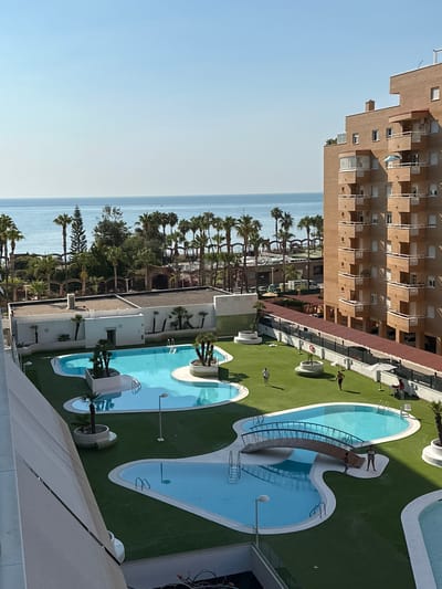 The image shows a bright, sunny day at a resort in Les Amplaries, Spain, with a view of the Mediterranean Sea. The foreground is dominated by a large, multi-level swimming pool complex with turquoise water and a decorative wooden bridge spanning one of the pools. The pools are surrounded by neatly manicured green artificial turf. Several palm trees and other tropical plants are scattered throughout the grounds. To the right, a tall, multi-story brick apartment building with numerous balconies overlooks the pool area. In the background, a line of palm trees and other vegetation separates the resort from the calm, blue sea. The sky is clear and a bright blue. Several people are visible in the image, including individuals walking on the grass, standing by the pool, and sitting at a table. One person appears to be working near the pool.