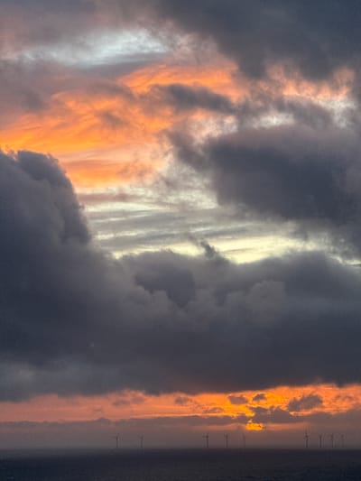 This media file captures a dramatic sunset or sunrise over a body of water, likely the sea, featuring a vast and expressive sky. The scene is dominated by a complex interplay of clouds, illuminated by the low-lying sun.
Large areas of the sky are filled with dark, heavy grey and bluish clouds, indicating a cloudy or potentially stormy atmosphere. These darker masses are interspersed with striking bands of light. In the upper-middle section of the sky, a prominent, elongated streak of bright, almost white-yellow light cuts horizontally across the frame, suggesting the sun's direct light piercing through or reflecting off higher clouds. Below this, closer to the horizon, another broad band of warm, intense orange and golden light illuminates the undersides and edges of numerous clouds, creating fiery textures and contrasts. Some clouds exhibit vertical streaking, catching the light in parallel lines.
Along the very low horizon, a series of dark silhouettes are visible against the orange glow: numerous offshore wind turbines, stretching across the distant seascape. This detail, combined with the location context, strongly suggests an offshore wind farm, possibly near Fécamp, France. The surface of the water beneath the turbines appears dark and calm.
No people are visible in the image, and there is no discernible text. The time of day is clearly the golden hour, and the overall mood is one of majestic natural beauty and powerful, dynamic weather conditions.