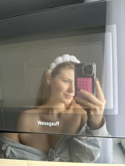 A close-up image depicts the reflection of an individual in the glass door of a "Weissgauff" branded microwave oven. The reflective surface features a perforated metallic mesh, which distorts the reflected image. The individual, appearing to be a female, wears a white, segmented headband and an unbuttoned light-colored shirt or robe with red piping, exposing her bare shoulders and upper chest. She is holding a smartphone, which has a prominent pink case, in her right hand, directing it towards the microwave door in an apparent act of taking a photograph of her reflection. Her expression is a subtle smile, with her eyes partially obscured by the mesh pattern. The background visible in the reflection is a plain, light-colored interior wall. The precise location within Tashevka, Russia, cannot be determined from the visual information presented.