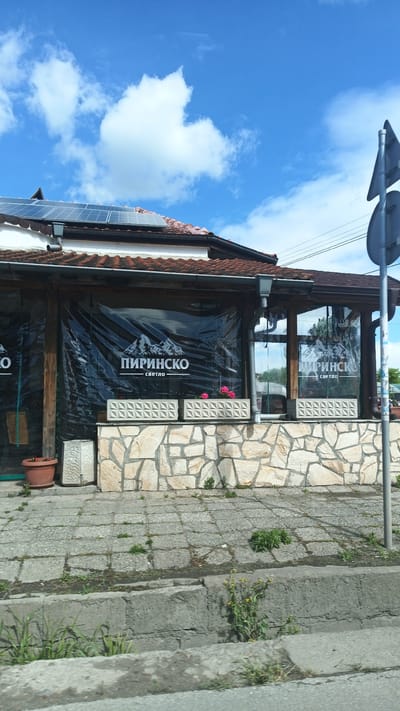 The image shows the exterior of a building in what appears to be Boliartsi, Bulgaria. The building has a visible sign reading "ПИРИНСКО" (PIRINSKO) with the word "светло" (svetlo) underneath, suggesting it is a restaurant or bar. The building features a tiled roof with solar panels, stone walls, and a covered patio area with transparent plastic sheeting bearing the PIRINSKO logo. The patio has concrete planters with pink flowers. The sidewalk in front of the building is paved with grey stones and shows some weeds growing in the cracks. The street is visible in the foreground, with a concrete curb. In the upper right corner, there is a glimpse of a road sign, suggesting it is located near a road intersection. The sky is blue with scattered white clouds.