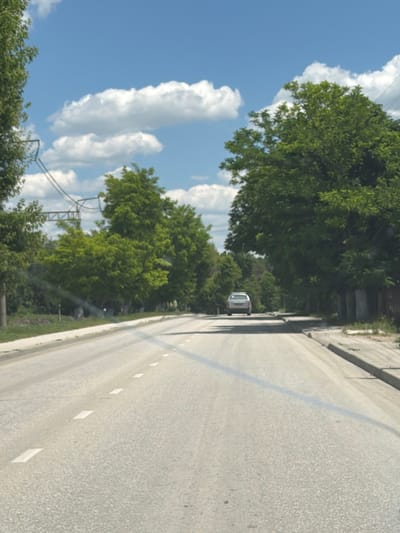 A car is driving down a paved road lined with trees on both sides. The sky is blue with some clouds. The road has white markings. There are power lines visible in the background. The environment appears to be a rural or suburban area, possibly in the city of Почтовское сельское поселение, Ukraine. The image is taken from a moving vehicle, as evidenced by the motion blur.
