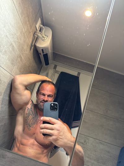 A man is lying on his right side on the tile floor in a bathroom. He is taking a selfie in the mirror. He is shirtless, revealing a muscular physique and tattoos on his chest and left arm. The location may be a private bathroom in Bredene, Belgium. The background includes a white wall-mounted heater, a grey tiled wall, and a dark colored garment hanging.