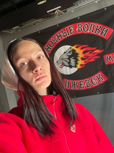 A woman wearing a red fleece jacket and a grey hoodie is posing for a selfie in front of a banner. The banner is predominantly black with a circular design featuring a wolf's head engulfed in flames in the center. Red Cyrillic text surrounds the central image. The location is Doma 45 Kilometr, Russia. The woman has brown hair, brown eyes, and multiple piercings in her nose.