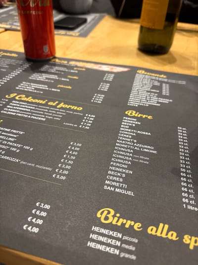 This close-up, high-angle shot depicts a dark-colored menu resting on a light wooden tabletop inside a restaurant in Foligno, Italy. The menu, featuring yellow text, displays food options such as "I Calzoni al forno" (baked calzones) and "Calzone fritto a piacere" (fried calzones), alongside sections for "Bevande" (drinks) and "Birre" (beers). Visible beer brands include Peroni, Heineken, Beck's, Bud, Moretti Rossa, Corona, Ceres, Tennent's, Nastro Azzurro, Ichnusa (also non filtrata and al limone), and San Miguel, listed with quantities like 33 cl, 50 cl, 66 cl, and 1 liter. Prices in Euros are visible for various items, ranging from €1.50 to €11.00. To the left, a red Coca-Cola Zero Caffeina aluminum can is partially visible. An out-of-focus dark green glass bottle, likely wine, is present on the right side of the table. A dark, indistinct rectangular object, possibly a smartphone, lies in the background. No people are visible.