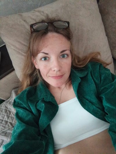 A close-up, overhead shot shows a woman lying down, propped up by a pillow. She is wearing a white ribbed tank top and a dark green corduroy button-up shirt. Her glasses are resting on top of her head. Her hair is a light brown and is spread out. She is looking directly at the camera with a slight smile. The background appears to be a couch or bed with a textured blanket. The lighting suggests it is daytime. No text is visible in the image.