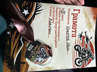 The image shows a certificate and a medal awarded for a motocross event. The certificate is light-colored with red and black graphics, including stylized flames and stars. Prominently displayed on the certificate is the word "Грамота" (Certificate) in large red letters, followed by "награждается Хребтов Иван" (awarded to Khrebtov Ivan). Below this, it states "За III место в Первом этапе Чемпионата и Первенства Удмуртской Республики по мотокроссу в классе Любители" (For 3rd place in the First stage of the Championship and Competition of the Udmurt Republic in motocross in the Amateur class). The medal is dark-colored with a graphic of a motocross rider. The medal also has text that is partially visible, including "2026" and "Трасса". The overall scene suggests an award ceremony or presentation of recognition for an achievement in motocross. The certificate and medal are likely on a table or surface, with a dark fabric visible on the left side of the frame. No specific location within a city is identifiable from the image itself.