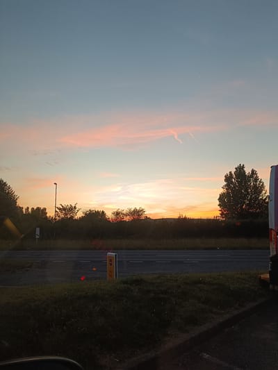 The image depicts a sunset scene, likely on a road in Arun, United Kingdom. The sky displays a gradient of colors, transitioning from a pale blue at the top to hues of orange and pink near the horizon. Wispy clouds are scattered across the sky.

In the foreground, a road is visible, with markings indicating lanes. To the right of the road, a grassy verge is present, and further beyond, a dark line suggests a ditch or embankment. On the left side of the image, a line of trees forms a dark silhouette against the brighter sky. A lamppost stands prominently in the middle ground, with a dark, leafless tree to its right.

Further to the right, another large tree with dark foliage is silhouetted. Part of a vehicle, appearing to be a white van with pink and white accents, is visible on the far right edge of the image. A yellow and white sign with the text "No Exit" is positioned near the road on the right side. The overall impression is of a roadside view at dusk.