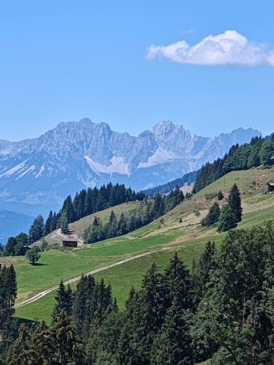 A scenic vista in Kochau, Austria, showcases a majestic mountain range in the distance, its peaks jagged and snow-dusted. The foreground is dominated by lush green hillsides dotted with coniferous trees. A winding dirt path traverses the landscape, leading to a rustic wooden building nestled amongst the greenery. The sky above is a clear, bright blue, with a solitary white cloud drifting lazily. The scene is tranquil and evokes a sense of natural beauty.