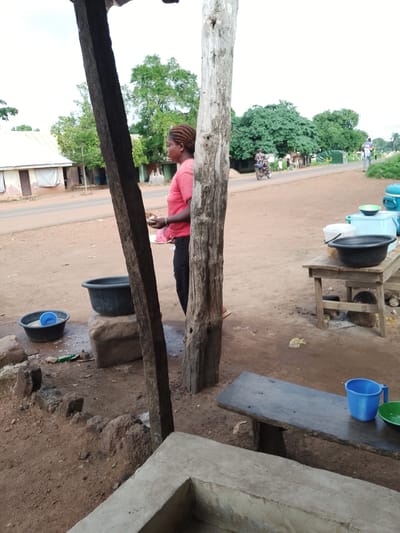 The image depicts an outdoor scene in Ichol, Nigeria, likely a roadside market or a small community gathering area. In the foreground, a woman with braided hair, wearing a pink t-shirt and black pants, stands between two large wooden posts, holding a plate with some food. To her left, two dark basins are visible on the ground, one containing a smaller blue cup and the other appearing to hold water. Further to the right, a wooden table is set up with several containers, including a large black basin, a white bucket, and a light blue cooler with a green bowl on top. A simple wooden bench is positioned in the lower right portion of the image, with a blue plastic cup and a green bowl placed on it. The ground is covered in reddish-brown dirt, characteristic of the Nigerian environment. In the background, a road runs horizontally across the frame, with buildings and lush green trees lining its edges. A motorcycle with a rider is seen on the road, and a few other figures are visible in the distance. The sky is overcast. The overall impression is of a casual, everyday setting in a rural or semi-urban area of Nigeria.