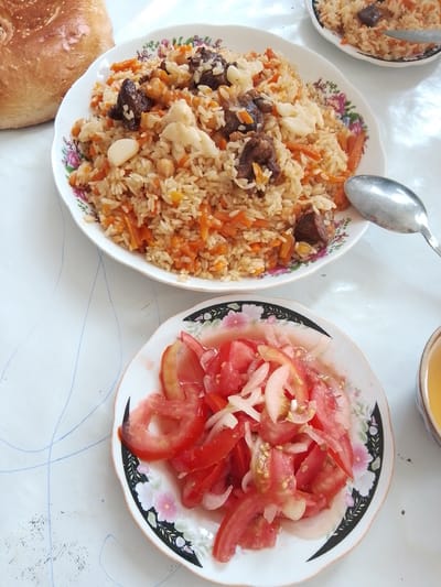 A top-down, slightly angled medium shot captures a meal laid out on a white tablecloth with faint blue lines. To the left, a golden-brown, round loaf of bread, likely flatbread, sits prominently. In the center, a white, floral-patterned plate holds a generous portion of pilaf, a rice dish mixed with carrots, chickpeas, and pieces of meat, with a clove of garlic visible. A silver spoon rests on the right side of this plate. Below the pilaf plate, another identical floral-patterned plate displays a salad of sliced tomatoes and onions. To the far right, a small portion of a cup with a golden liquid, possibly tea, is visible. The scene depicts a traditional meal likely served in Jizzakh, Uzbekistan.