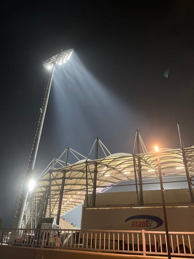 A nighttime view captures a stadium's exterior, with bright floodlights illuminating the dark sky and generating a smoky effect. Metal scaffolding supports the lighting structure. A stadium roof, adorned with "azamtv" branding, has a curved, fabric-like covering supported by a network of metal beams. A section of stadium seating appears in the background. The image is taken from a low angle.