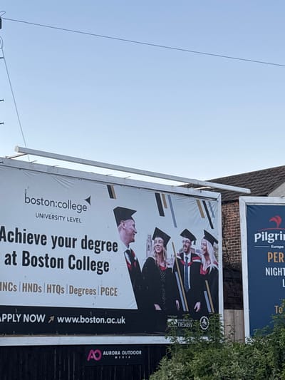 The image shows a billboard for Boston College, located in Boston, United Kingdom. The billboard features the Boston College logo and the tagline "Achieve your degree at Boston College". Below this, it lists various qualifications such as HNCs, HNDs, HTQs, Degrees, and PGCE. The bottom of the billboard displays "APPLY NOW" and the website address "www.boston.ac.uk". The billboard also shows images of graduates wearing academic regalia. To the right of the Boston College billboard, a portion of another billboard for "Pilgrim" is visible. In the foreground, lush green foliage obscures the lower part of the billboards. The sky is clear and light blue, suggesting daytime.