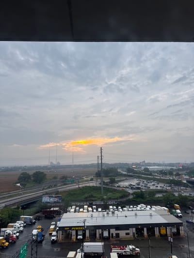 The image depicts an overcast sky with a sliver of yellow sunlight breaking through the clouds, situated above a sprawling industrial and commercial area in North Bergen, United States of America. The foreground is dominated by a large Penske truck rental facility with numerous trucks parked in its lot. Several other trucks, including box trucks and tow trucks, are visible throughout the scene. In the middle ground, a multi-lane highway system curves through the landscape, with vehicles in motion. Beyond the highway, a marshy area borders a body of water. In the distance, tall radio towers and city buildings are faintly visible through the hazy atmosphere. The overall impression is one of an early morning or late afternoon industrial scene under a dramatic sky.