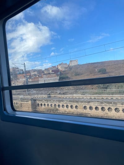 The image displays an exterior view from a train window, looking out over a landscape in Sidi Kacem, Morocco, on a clear day with a blue sky and scattered white clouds. A prominent hillside dominates the background, featuring a cluster of multi-story buildings constructed from brick and concrete on its lower slope to the left, many appearing unfinished or in various states of repair. Further up the hill to the right, isolated light-colored structures are visible. Utility poles and overhead lines run parallel to the train tracks in the mid-ground. The foreground reveals a railway line with at least two parallel tracks. Directly beneath the tracks is a concrete or stone embankment, possibly part of a bridge structure, adorned with a repeating pattern of recessed octagonal or star-shaped openings. Sparse, dry vegetation is present on the hillside and adjacent to the railway embankment. No individuals are depicted in the scene.