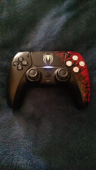 A matte black PlayStation 5 DualSense controller is centrally positioned, resting on a dark, textured fabric surface, likely a blanket or carpet. The controller features a white Spider-Man emblem above the central PlayStation button. The right side of the controller, including the face buttons and grip, is customized with a red and black pattern resembling organic branches or webs. The standard white PlayStation face buttons (Triangle, Circle, Cross, Square) are visible on the right. The directional pad buttons on the left are black. A blue light bar is illuminated above the PlayStation button, indicating the controller is powered on or charging. No individuals, actions, or interactions are present. The environmental lighting is low. The precise location within the city of Torreón, Mexico, cannot be determined from the visual information provided.