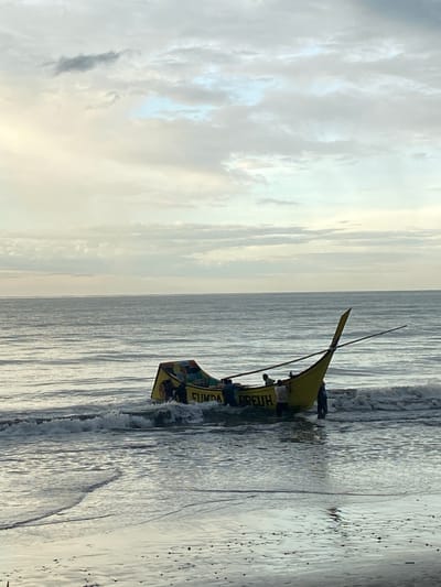 The image depicts a yellow fishing boat with the words "FUNDA PREUH" written on its side, partially submerged in shallow ocean water near the shore of Bateetimoh, Indonesia. Several individuals, appearing to be fishermen, are visible on and around the boat, actively engaged in what seems to be launching or docking operations. The water is calm with small waves breaking around the boat. The sky above is overcast with a mix of gray and light blue clouds. The overall scene suggests a typical day for fishermen in a coastal Indonesian town.