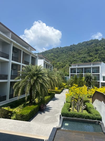The image shows a hotel complex situated in Baan Klong Prao, Thailand. The complex consists of multiple multi-story, white buildings with balconies, interspersed with tropical vegetation. Palm trees are a prominent feature, along with flowering trees and manicured shrubbery. A mountain covered in dense forest forms the backdrop. A paved walkway runs through the complex, leading to a water feature that appears like a small, rectangular pool. The sky is blue with a scattering of clouds. There are no people visible in the image.