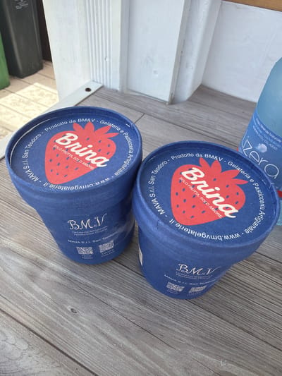 Two blue ice cream cups with red strawberry graphics sit on a weathered wooden surface, likely a table or boardwalk, in Santu Diadòru (San Teodoro), Italy. The cups are branded "Brina" and feature text in Italian indicating they are from "BM&V - Gelateria e Pasticceria Artigianale," with a website address "www.bmvgelaterie.it" and "MAVA S.r.l. San Teodoro." One cup is slightly to the left and in the foreground, while the other is to the right and further back. Partially visible in the background is a blue plastic bottle of mineral water with Italian text. The scene suggests an outdoor setting, possibly near a shop or kiosk.