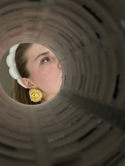 A face peers through the circular opening of a gray tube. The visible eye is light with sparse lashes. A golden, textured earring adorned with small diamonds is on the visible ear. A white, fluffy headband sits on the hair. The tube walls are textured, appearing to be a tunnel of printed material. The lighting is soft, casting shadows and highlighting the textures.