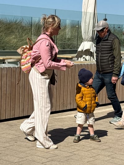 The image captures three individuals on an outdoor wooden promenade or deck in Middelkerke, Belgium, under bright, clear daylight.
In the foreground, an adult woman is visible in profile, facing left. She has blonde hair tied back and is wearing a pink jacket, wide-leg white pants with thin vertical stripes, and white sneakers. A colorful, striped backpack is slung over her shoulder. She appears to be looking down at a smartphone she holds in her hands.
Behind her, slightly to the right, a young child stands facing upwards, looking towards the man. The child is dressed in a bright yellow puffer jacket, a dark blue beanie, white shorts, and green sneakers with white laces. Strong shadows are cast from the child onto the paved ground to their right.
Further back, an adult man is walking to the left, partially obscured by the child and a large folded white umbrella. He wears a grey baseball cap, a dark puffer vest over a dark long-sleeved shirt, and blue jeans. His right hand is visible, possibly holding something or gesturing.
The setting includes a wooden deck underfoot, transitioning to a light-colored paved area on the right. In the background, there's a glass balustrade, beyond which green dune-like vegetation is visible under a clear blue sky. The strong sunlight casts distinct shadows, indicating a bright, sunny day, likely mid-day or afternoon. No visible text is present in the image. The overall scene depicts a casual outdoor stroll.