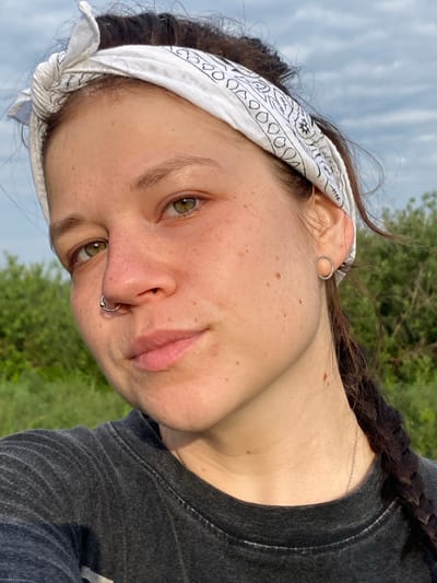 A close-up, eye-level, outdoor selfie shows a young woman from the chest up. She is looking at the camera with a neutral expression. Her hair is dark brown and pulled back with a white bandana patterned with black paisley designs. She has green eyes, freckles across her nose and cheeks, and a septum piercing. She is wearing a dark grey t-shirt. The background is blurred green foliage and a cloudy sky. The lighting suggests it is daytime. The location is not specified in the image.