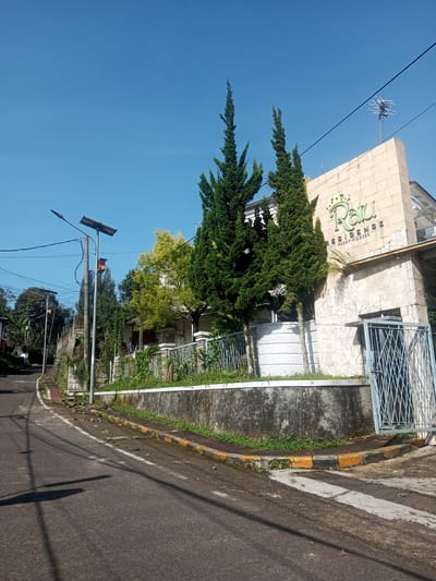 The photograph shows a street scene under a clear blue sky. On the right is the entrance to "Ratu Residence, Passir Cianjur," marked by a beige stone building with a gate. Two tall, slender evergreen trees stand in front of the building.

Along the left side of the image, a road curves slightly uphill. A street lamp with a solar panel stands adjacent to a concrete retaining wall that runs along the roadside, above which are trees and foliage.

The road surface is asphalt, showing signs of wear and patching. The curb is painted with alternating yellow and black sections. Small patches of vegetation grow in the spaces between the curb and the road. There are no people visible in the image. The photograph does not show any location in Nhot Chang, Laos.