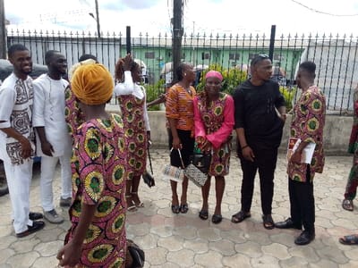 The image shows a group of people standing outside on a paved sidewalk in front of a black metal fence. The scene is set in Yenagoa, Nigeria at the coordinates 4.931 latitude and 6.267 longitude. There are seven people in the photo, all of whom are adults. Four are wearing clothing with patterned designs. The two in the front, one with a yellow hat and the other with a black shirt, are facing forward. They are both holding a bag. In the back, a woman in a pink shirt is speaking with a man in a black shirt. The other individuals appear to be listening to them. The people are standing on a pavement made of hexagonal blocks and behind the fence is a building with a green wall.