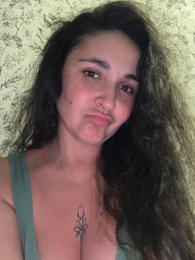A self-portrait of a woman with dark, long, curly hair and brown eyes, wearing a green tank top. She has a tattoo on her chest. The background is a light green wallpaper with a floral pattern. The woman is making a kissing face. The image was taken indoors.