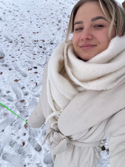 An overhead-angle portrait depicts a young adult female outdoors in Доња Брезна, Montenegro, during winter. The subject occupies the right half of the frame, facing the camera with a slight smile. She has light-colored hair, dark eyebrows, and is wearing a cream-colored puffer coat cinched at the waist with a tie-belt, and a thick, cream-colored knitted scarf wrapped around her neck. The ground is covered in fresh snow, interspersed with numerous brown, dried leaves and multiple distinct boot or shoe prints. A neon-green cord, possibly a leash, with a black cylindrical connector at its end, extends from the lower-left foreground towards the subject's right arm, which is outside the frame. A faint brown object, potentially a wooden bench, is partially visible in the upper right background, behind the subject's head.
