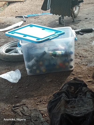 The image shows a clear plastic bin with a blue lid, containing various bottles and other items. The bin is located on a dirt surface with a tire in the background. The blue lid of the bin is open, revealing the contents. There is a black cloth or tarp in the lower right corner of the image. The scene appears to be an outdoor area in a town or village setting. The location is in Aramoko, Nigeria.
