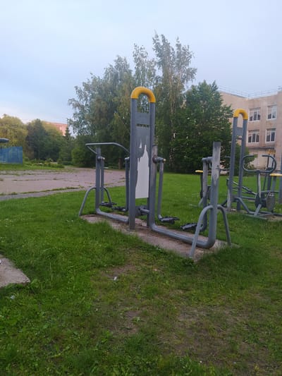 An outdoor exercise equipment area is depicted, situated in a grassy, somewhat unkempt park setting with a paved area visible in the background. The sky is overcast and a soft, diffused light illuminates the scene. In the foreground, two grey metal exercise machines are prominent. The machine on the left features a vertical support with horizontal bars and foot pedals, designed for leg movement. The machine on the right is partially visible and appears to be a different type of exercise apparatus.
Behind the exercise equipment, a large, multi-story building with a light-colored facade and numerous windows dominates the background. The building is positioned to the right of the frame. Trees with green foliage are scattered throughout the scene, some taller and fuller, others more sparse. A blue fence is visible in the far left background, suggesting the edge of the park or a neighboring area. The ground is covered with green grass, with patches of bare earth and gravel interspersed, indicating a lack of consistent maintenance. The overall impression is of a public outdoor space that has seen better days. The location is identified as Gusev, Russia.