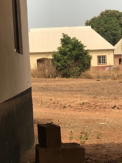 The image depicts an outdoor scene in Egboje, Nigeria, under a hazy, light grey sky. In the immediate foreground on the left, a cream-colored building wall with a dark grey lower section partially obscures the view. Two stacked, hollow, weathered concrete blocks are positioned at the base of this wall, with a small, dark lizard or similar reptile visible on the upper block.
The midground consists of a wide expanse of reddish-brown earth, sparsely covered with dry, light-colored grass and scattered small green plants. The ground surface shows minor undulations and faint linear disturbances.
In the background, a single-story building with light yellow walls and a light-colored, sloped corrugated metal roof is centrally located. An arched, dark opening is visible on the left side of this building. A dense, green-leafed tree stands directly in front of the building's central portion. To the right of the tree, a rectangular window with white frames and multiple panes, surrounded by a reddish-brown trim, is set into the yellow wall. A section of another building, featuring a similar light-colored roof and reddish-brown lower structure, is partially visible on the far right. Taller, darker green trees are discernible behind the structures in the distant background. No individuals or discernible activities are present in the scene.
