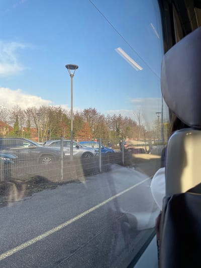 The image depicts an exterior scene in Dettwiller, France, viewed from inside a vehicle, likely a train or bus, through a window. The upper frame is dominated by a clear blue sky with sparse white clouds. Below the sky, a line of deciduous trees with mostly bare branches, and some with reddish-brown foliage, forms the horizon. Residential buildings with orange/red roofs are partially visible behind the trees on the far left.
In the midground, a dark grey, modern streetlamp with a flat, circular luminaire extends vertically. To its right, a metal grid fence runs horizontally, separating an asphalt road or pathway in the immediate foreground from a gravel parking area. A solid yellow line is painted on the asphalt surface.
Within the parking area, multiple vehicles are parked: a dark-colored SUV (partially visible on the left), a silver/grey SUV, a blue car, and a brown/bronze SUV (visible on the right). Two adult male figures, dressed in dark attire, are standing near the blue and brown SUVs, in proximity to a white and red railway level crossing barrier that is partially visible on the far right.
The vehicle window displays reflections, including a long, rectangular light source in the upper right quadrant, a light grey/white vehicle seat and a purplish headrest on the far right, and a faint human silhouette. The scene is illuminated by bright daylight, casting visible shadows.