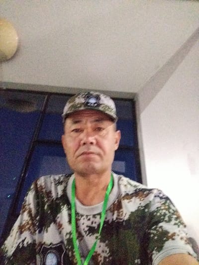 A man wearing a camouflage hat and shirt with a green lanyard around his neck is pictured. He is looking directly at the camera with a neutral expression. The background consists of a window with dark blue panes and a portion of a white ceiling with a circular light fixture. The scene appears to be indoors, possibly in a room with large windows. The lighting suggests it might be daytime, though the indoor setting makes it difficult to determine the exact time of day or weather conditions. The man's attire, particularly the camouflage pattern, could imply an outdoor or recreational activity. There is no visible text in the image. The provided location context, "Түндүк айланып өтүүчү жол 84 га, Kyrgyzstan," suggests the photo may have been taken in Kyrgyzstan, along a northern bypass road.