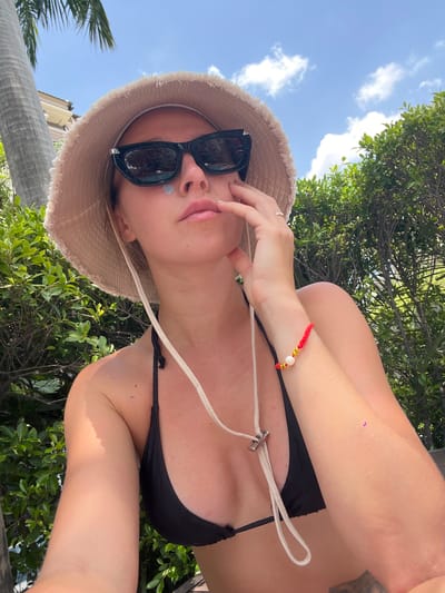 A woman is taking a selfie outdoors, wearing a black bikini top, a wide-brimmed hat, and sunglasses. She has a red bracelet with beads on her left wrist. Behind her, there is lush green foliage and a bright blue sky with scattered clouds. The scene appears to be a resort or a tropical location, possibly Bang Sao Thong Subdistrict, Thailand, given the presence of palm trees and the overall ambiance.
