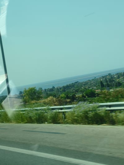 The image captures a scenic view from a moving vehicle, likely a car, looking towards the sea. The foreground shows a paved road with a white line marking a lane. A metal guardrail runs along the side of the road, separating it from a lush green embankment covered with grass and some flowering plants.

Beyond the embankment, a hillside is visible, dotted with numerous houses and buildings with red-tiled roofs, indicating a residential area. The houses are nestled amongst dense green trees and vegetation, creating a picturesque landscape. In the distance, a vast expanse of calm, blue water stretches to the horizon, suggesting the presence of a sea or large lake. The sky is a clear, light blue, with a faint, elongated shadow or smudge visible high up, possibly a contrail or a smudge on the window.

The lighting suggests it is daytime, with the sun likely overhead, casting soft shadows. The overall impression is one of a tranquil, coastal setting, characteristic of many towns and villages in Greece, specifically Nea Kallikrateia, known for its proximity to the sea and its hillside residential areas. There are no people or animals clearly visible in the frame. The image seems to be taken from a slightly elevated position, consistent with being on a road that overlooks the coastline.