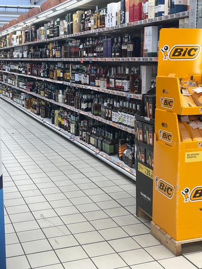 The image shows a supermarket aisle with shelves stocked with various alcoholic beverages. In the foreground, there is a bright yellow display stand with "BiC" branding, promoting what appear to be lighters or stationery products. The shelves are filled with bottles of wine, spirits, and other liquors. The flooring is tiled with a white and grey checkered pattern. In the background, other aisles of the supermarket are visible. The scene is well-lit, typical of a retail environment. The specific location in Castiglione delle Stiviere, Italy, is not evident from the image itself but is provided as context. There are no people visible in the image.